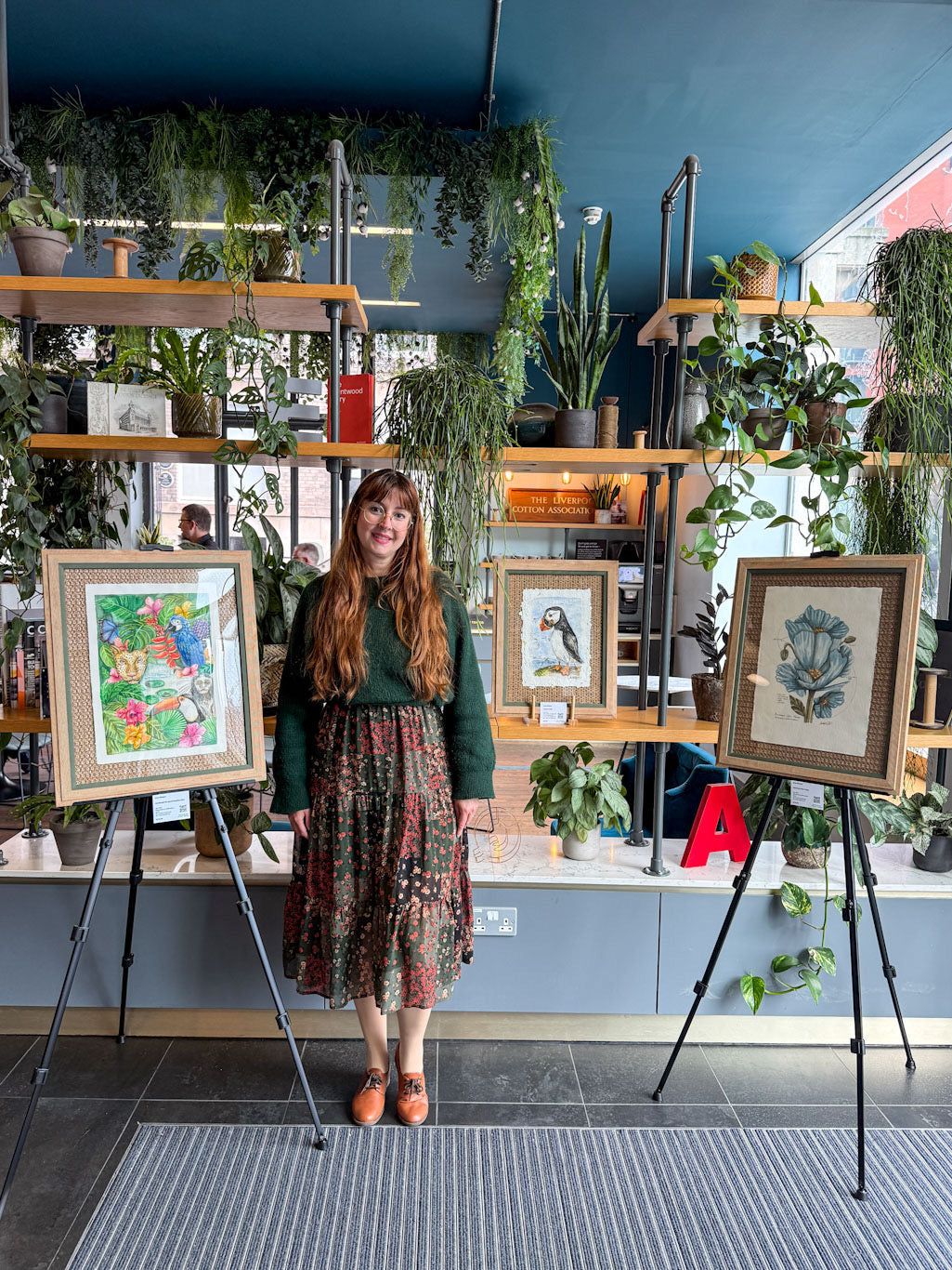 Woman standing in a room with art on easels and plants on shelves.