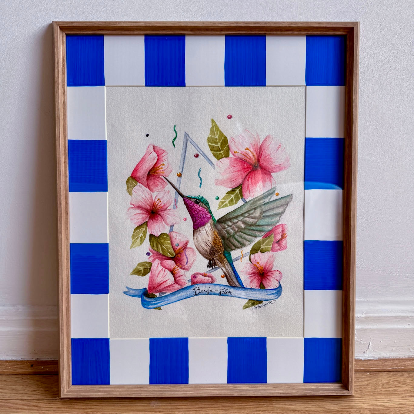 Framed artwork of a hummingbird and flowers on a white background with blue squares, against a white  wall.